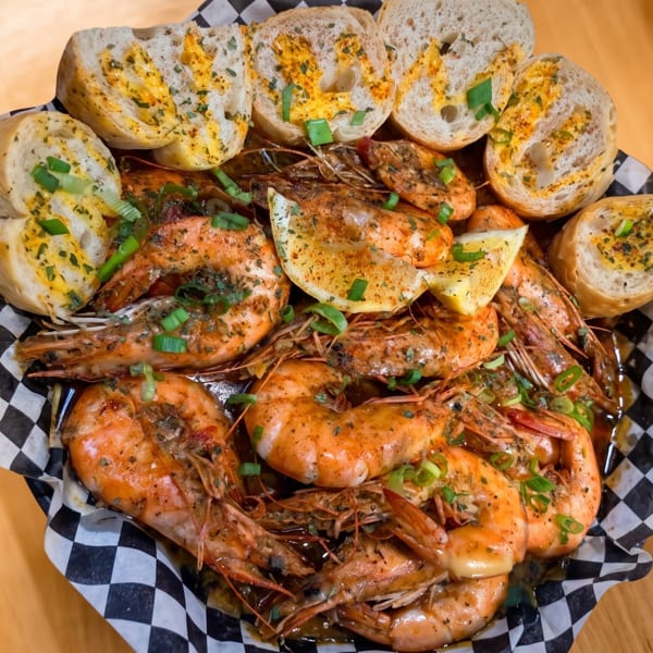 A basket of seasoned shrimp with lemon slices and garlic bread on checkered paper.